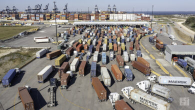 Lines of trucks queue up at a port terminal with cranes in the background.