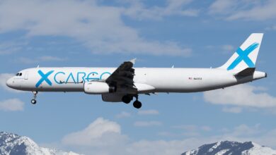 A GlobalX freighter with light-blue markings on airport approach with wheels down, blue sky in background.