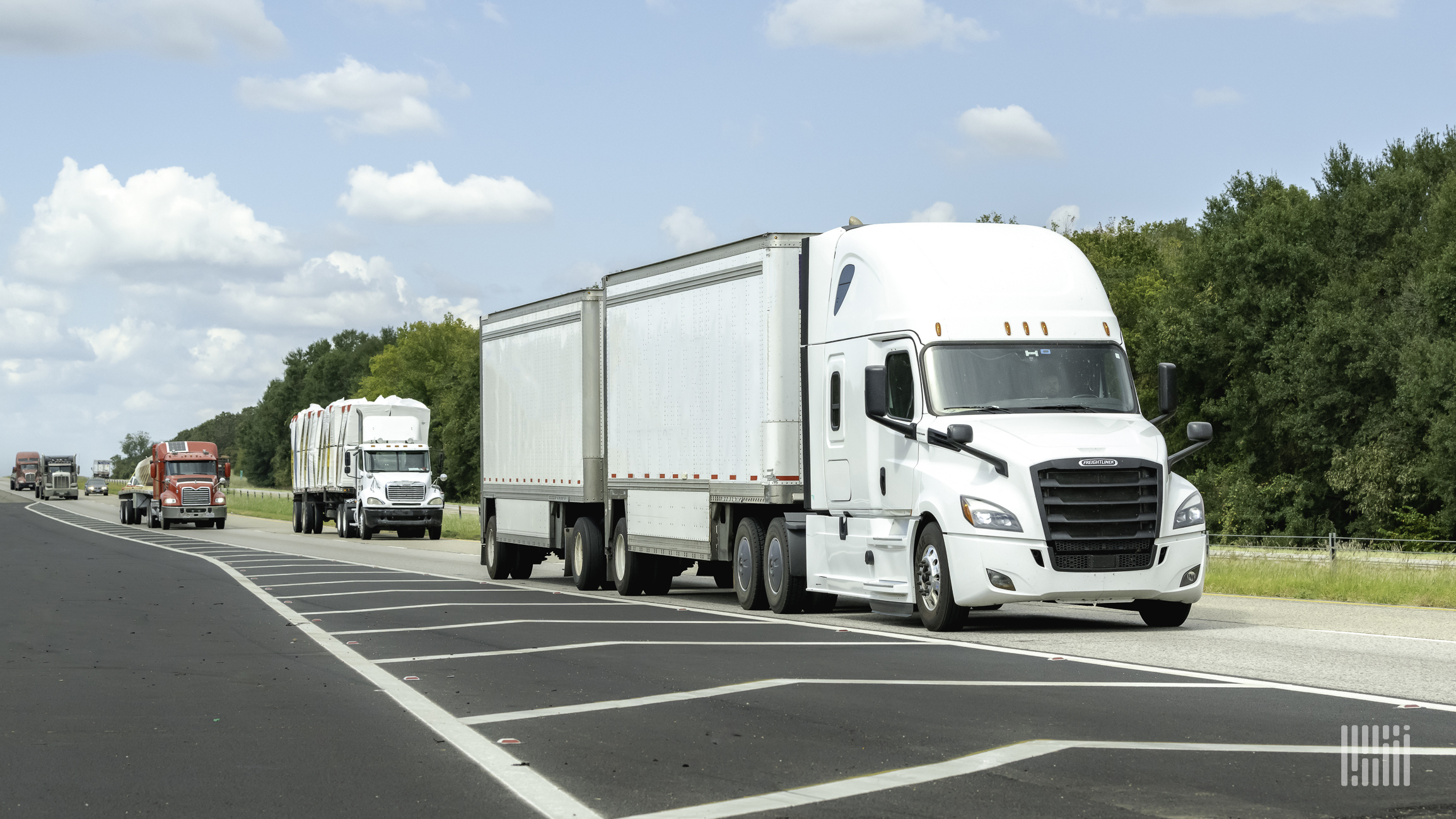 A white semi truck is followed by other trucks on a highway with green trees in the background.